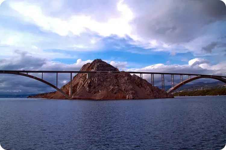 Blick vom Meer auf die Krk-Brücke mit markanter Felskuppe in der Mitte – dramatischer Wolkenhimmel über der Adria in der Nähe von Camping Omišalj, Insel Krk, Kroatien