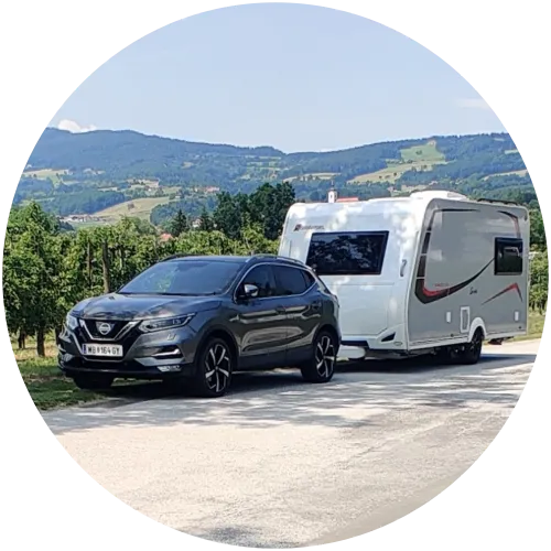 Nissan Qashqai zieht einen weißen Wohnwagen auf einem Parkplatz inmitten von Weinbergen und grüner Hügellandschaft in Österreich