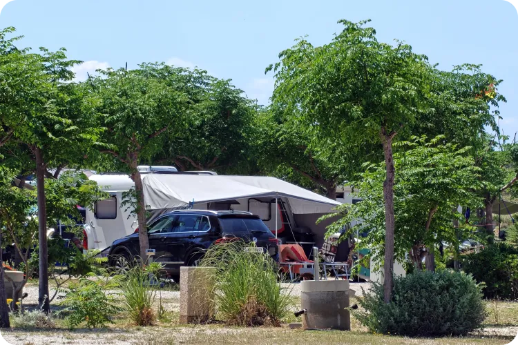 Ein Stellplatz auf dem Campingplatz Omišalj mit einem geparkten Wohnwagen, einem davorstehenden schwarzen Pkw und einem ausgefahrenen Vorzelt, umgeben von schattenspendenden Bäumen.