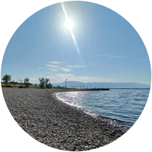 Sonniger Kieselstrand und blaues Meer am Camping Omišalj Ein geschwungener, heller Kieselstrand am klaren, blauen Meer des Campingplatzes Omišalj unter strahlender Sonne und blauem Himmel mit Bergen im Hintergrund.