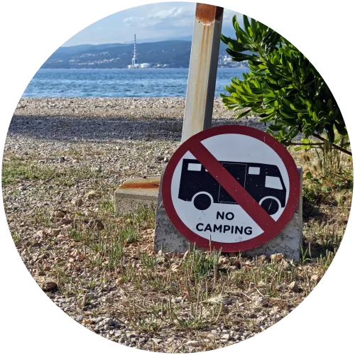 Camping-Verbotsschild an der Küste von Omišalj Ein rundes Verbotsschild mit der Aufschrift „No Camping“ und einem durchgestrichenen Wohnmobil-Symbol steht auf kieseligem Boden an der Küste von Omišalj mit Blick auf das Meer.