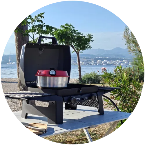 Kochen mit dem Omnia Backofen auf dem Enders Gasgrill am Meer Ein roter Omnia Campingbackofen steht auf einem Enders Urban Pro Gasgrill, der auf einem Campingtisch direkt am Strand des Campingplatzes Omišalj aufgebaut ist, mit Blick auf das Meer und die Berge.