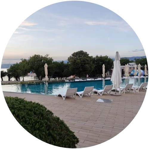 Großzügiger Swimmingpool mit Sonnenliegen am Camping Omišalj Ein großer, blau schimmernder Swimmingpool auf dem Campingplatz Omišalj, umgeben von weißen Sonnenliegen, geschlossenen Sonnenschirmen, grünen Bäumen und mit Blick auf das nahe Meer im Hintergrund.