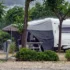 Weißer Wohnwagen mit grauem Vorzelt steht bei starkem Regen auf schattigem Campingplatz zwischen Bäumen