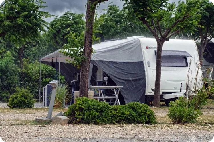 Weißer Wohnwagen mit grauem Vorzelt steht bei starkem Regen auf schattigem Campingplatz zwischen Bäumen